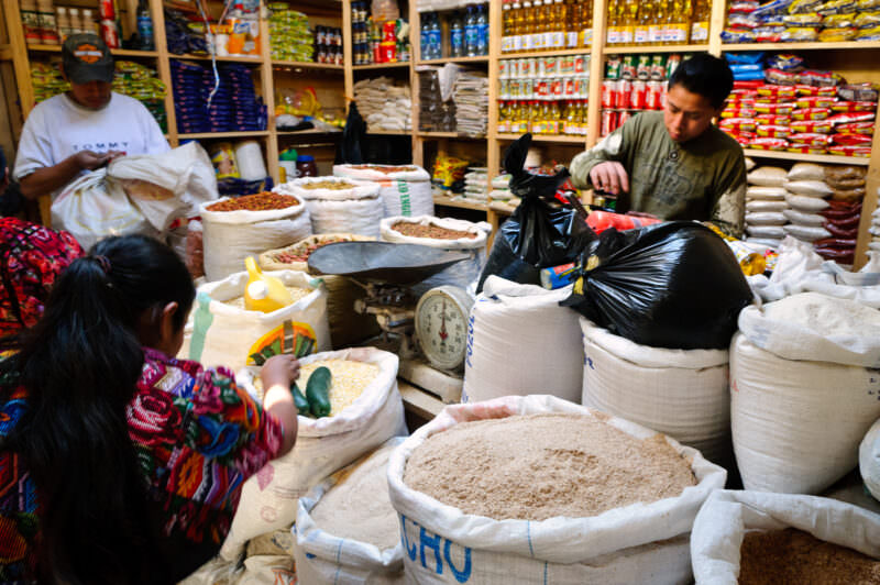 Market vendors beside open sacks of grains and seeds on wooden floor, shelves of packaged goods behind
