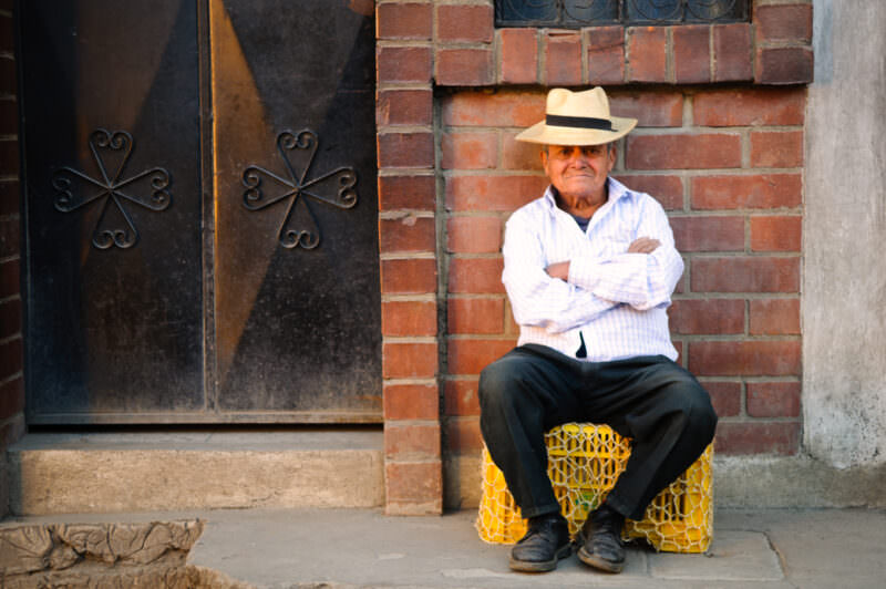 Man in white straw hat and white shirt seated on yellow plastic crate against red brick wall and ironwork door