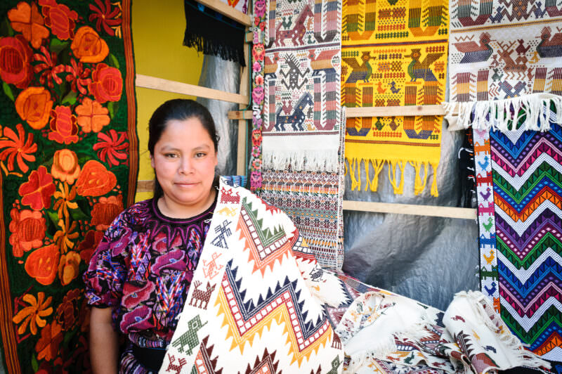 Woman in traditional purple embroidered huipil holding cream-colored woven textile, patterned textiles hanging behind