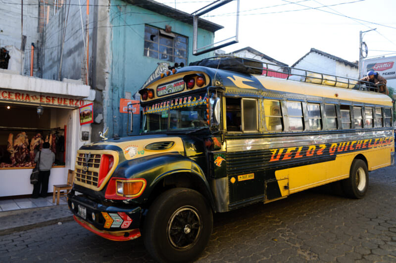 Yellow chicken bus with decorative stripes and lettering navigating cobblestone street in Chichicastenango