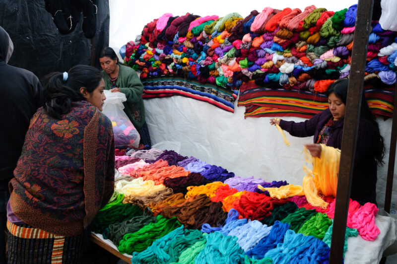 Market stall displaying hundreds of colorful thread skeins in rainbow arrangement for weaving, vendors browsing