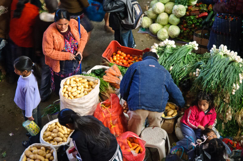 Woman in orange shawl beside baskets of potatoes and carrots, children seated nearby in vegetable market