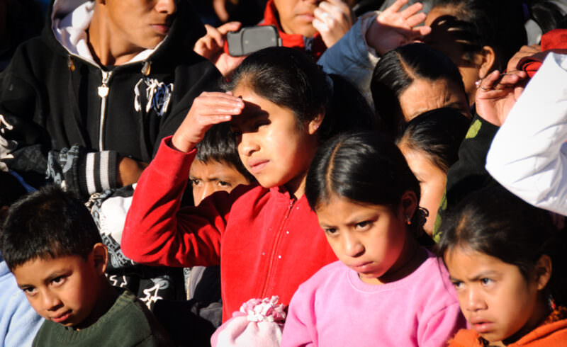 Row of children in bright jackets shielding eyes from sun, watching performance in crowd