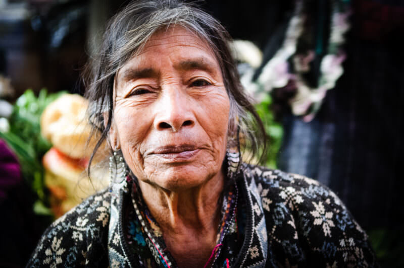 Elderly Maya woman with silver hair and embroidered black blouse, direct gaze, flower vendor stalls behind