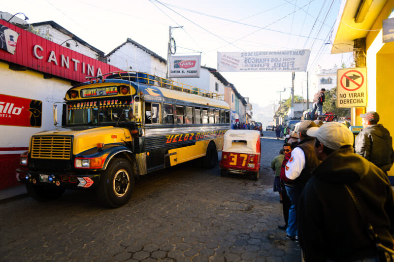 Yellow chicken buses and pedestrians on cobblestone street under tangled power lines, storefronts and cantina signs visible