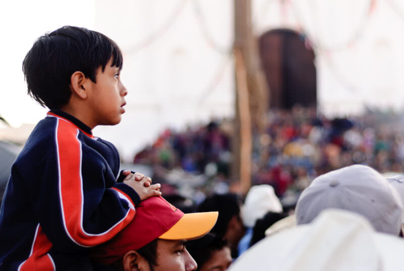 Boy in navy blue jacket sitting on adult's shoulders, profile view against blurred crowd at festival