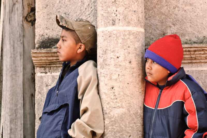 Two boys in profile leaning against weathered columns, one in tan cap, one in red knit cap