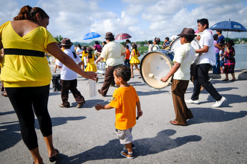 Young boy in an orange shirt holding hands with a woman as a marching band with drums passes by in the Independence Day parade
