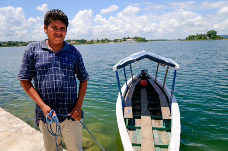 Boatman in a blue checkered shirt standing at the shore beside his traditional covered wooden boat on Lake Petén Itzá