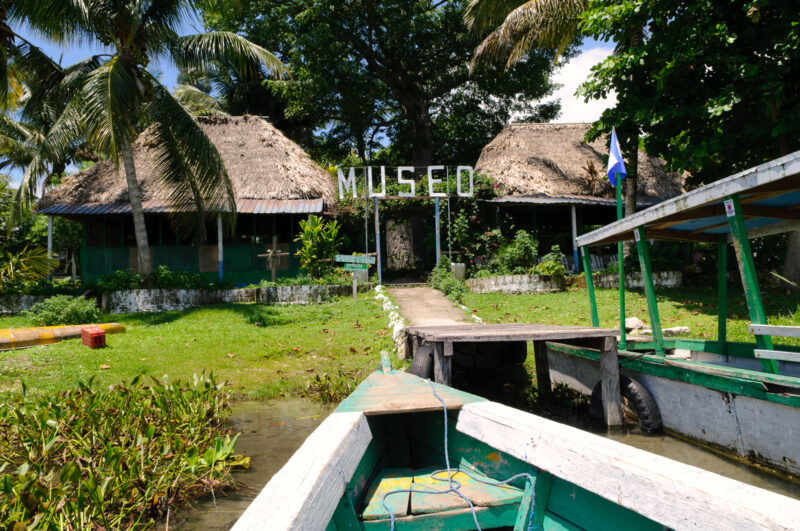 Thatched-roof museum buildings on Radio Petén Island with a Guatemalan flag, approached by a small green boat moored at a wooden dock
