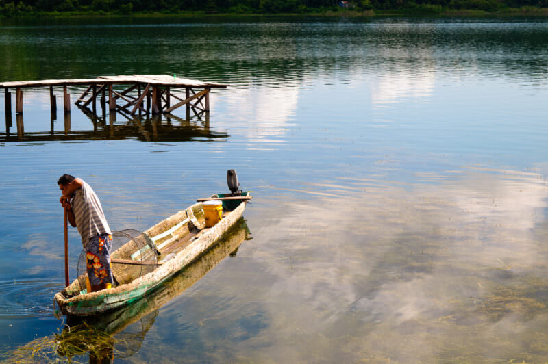 Fisherman wading beside a traditional wooden boat in the shallow, mirror-like waters of Lake Petén Itzá near Flores