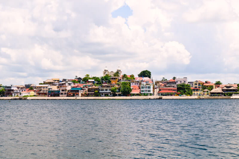 Flores island town viewed from Lake Petén Itzá, showing colorful multi-story buildings clustered along the waterfront under cloudy skies