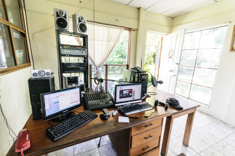 Radio Petén studio interior showing mixing desk, computers, speakers mounted on the wall, and windows overlooking the island's greenery