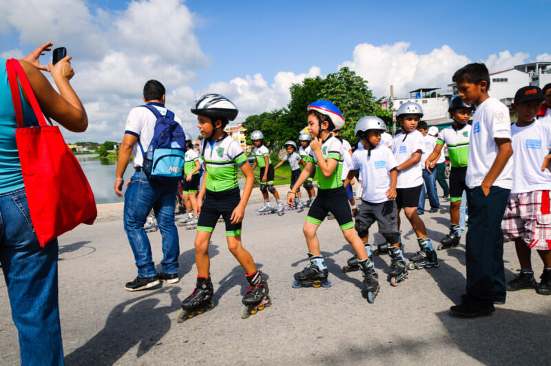 Boys on rollerblades in white and green uniforms skating in the Guatemalan Independence Day parade, with spectators and Lake Petén Itzá in the background
