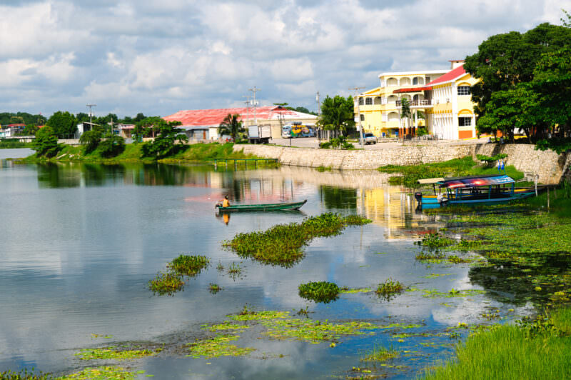 Colorful lakeside buildings in Flores reflected in the calm waters of Lake Petén Itzá, with small boats and floating vegetation in the foreground