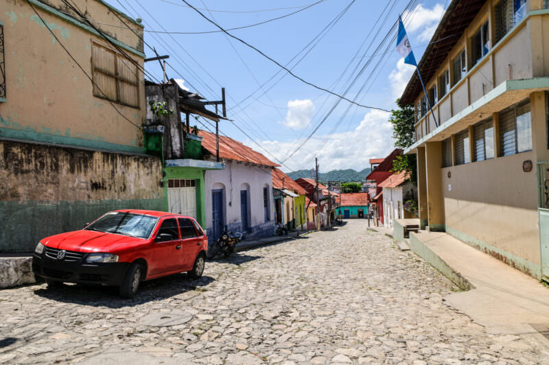 Cobblestone street in Flores, Guatemala, lined with colorful colonial buildings and a red Volkswagen parked on the left under overhead power lines