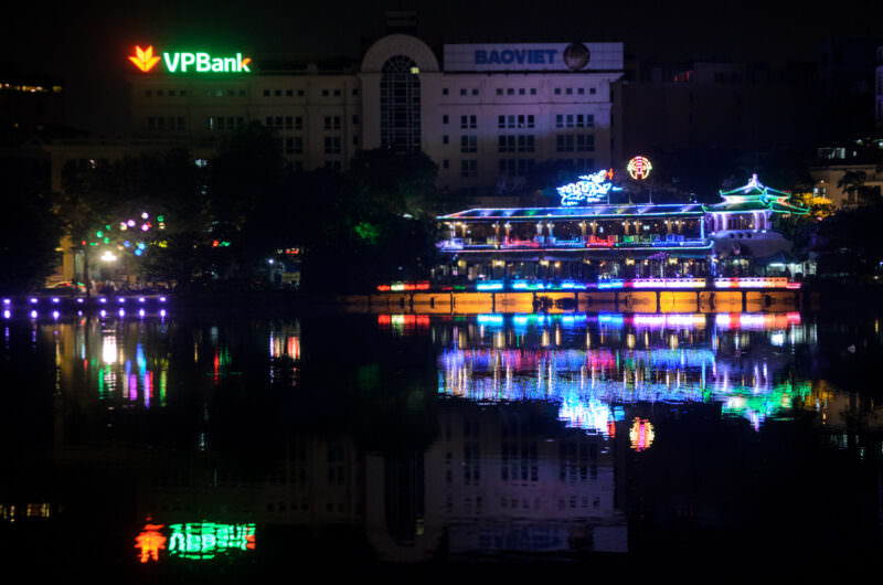 Neon signage and the illuminated Thuy Ta Cafe pavilion reflected across Hoan Kiem Lake at night