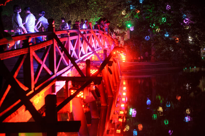 Pedestrians crossing the red wooden trusses of The Huc Bridge toward Jade Island at night