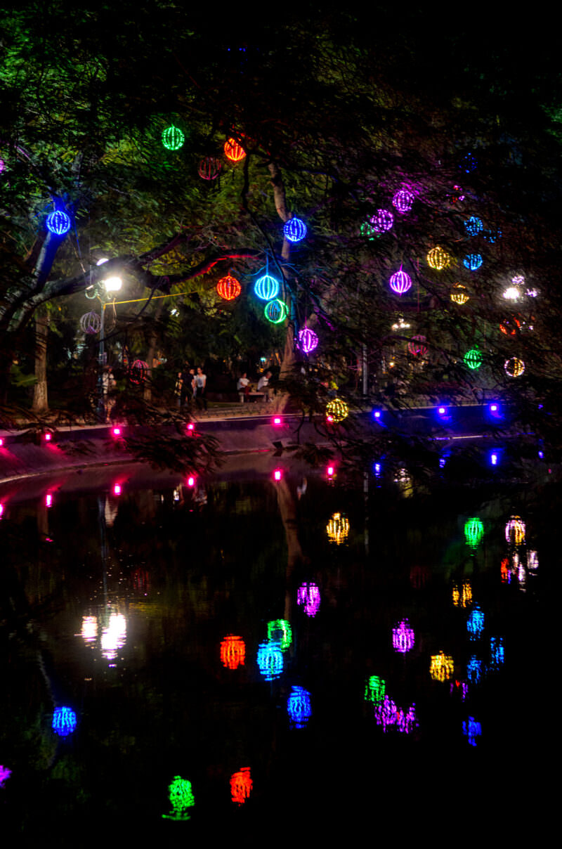 Colorful lantern lights hanging over trees reflected in the dark water of Hoan Kiem Lake