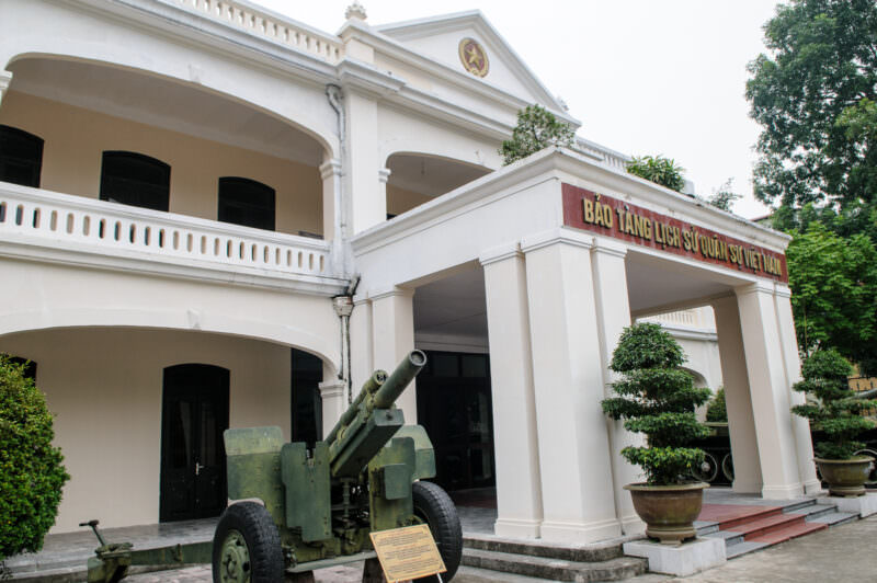 Artillery piece parked before the colonnaded neoclassical facade of the Vietnam Military History Museum