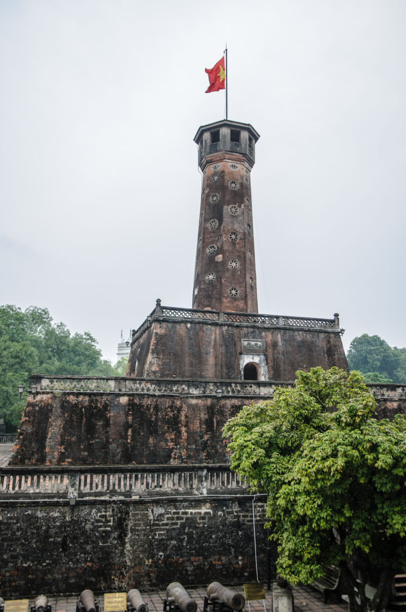 The tall octagonal Hanoi Flag Tower with red flag flying above a row of old cannons