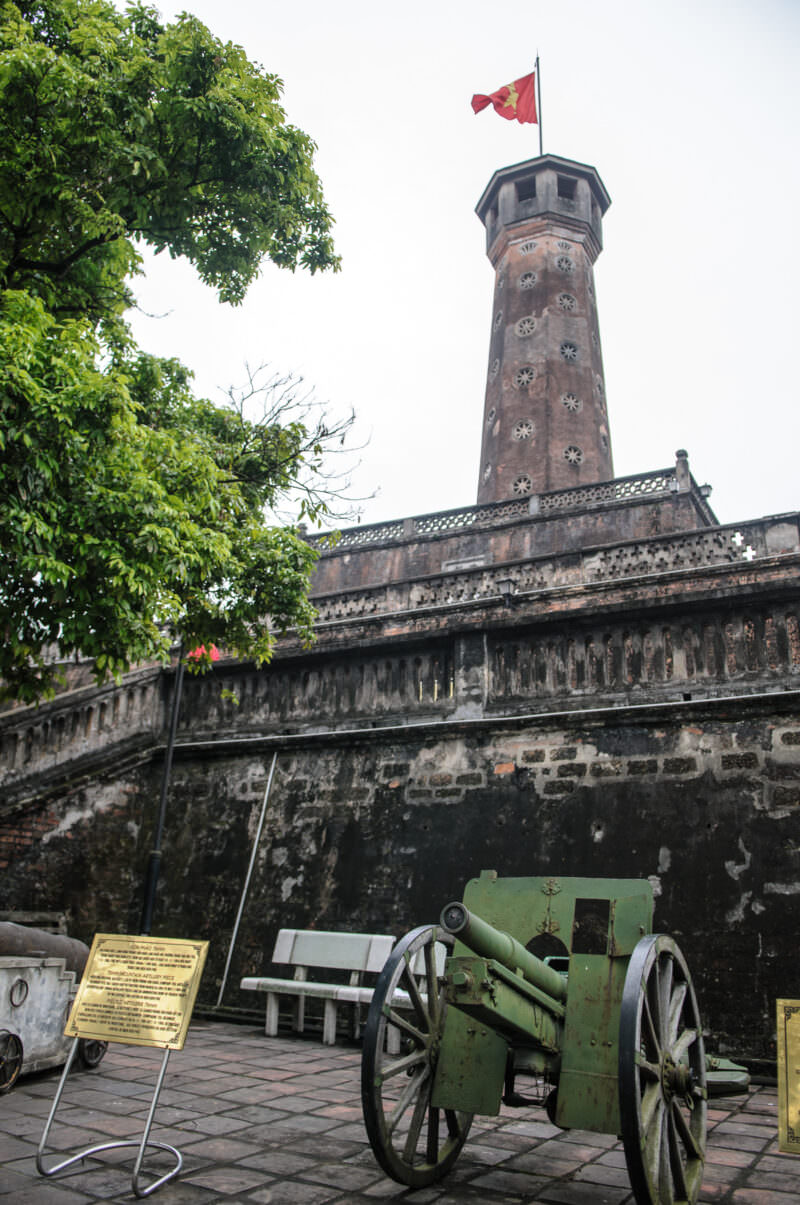 A green wheeled mountain artillery gun standing before the tall brick Hanoi Flag Tower