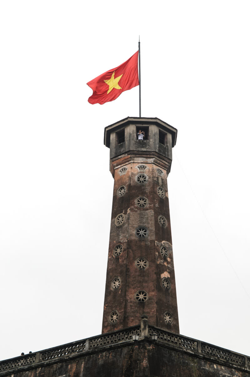Hanoi Flag Tower's octagonal brick shaft topped with the Vietnamese flag against an overcast sky