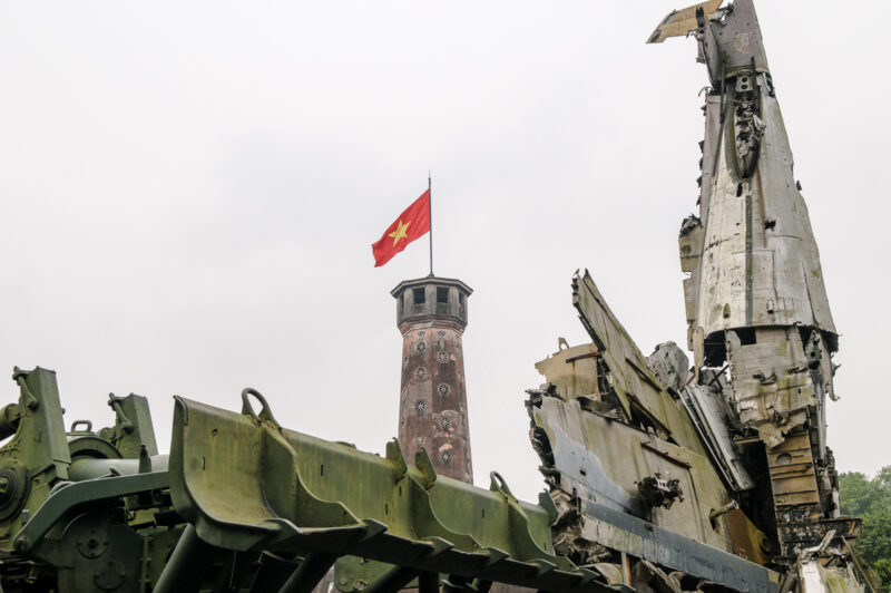 Mangled downed aircraft wreckage in the foreground with the Hanoi Flag Tower rising behind it