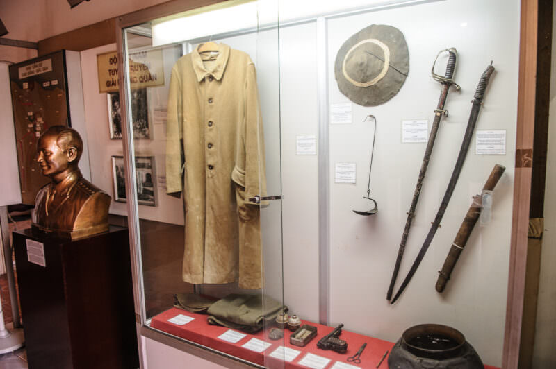 Glass case displaying a Viet Minh overcoat, machete, and sword alongside a bronze bust