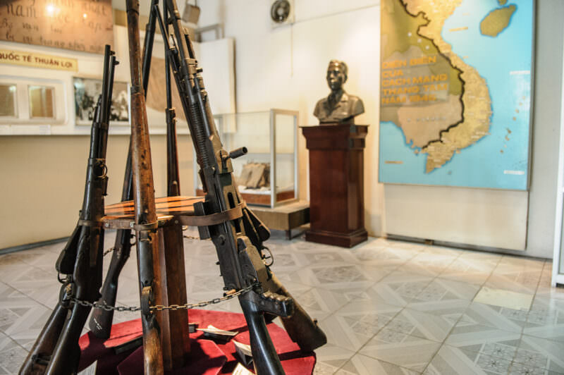 Chained vintage rifles and submachine guns displayed before a bust and Vietnam map in a museum gallery