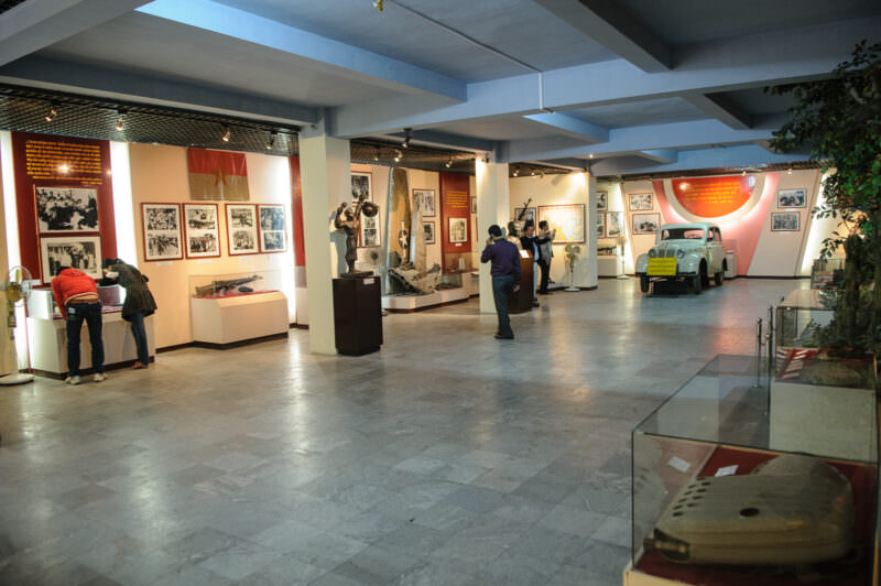 Museum gallery with visitors viewing aircraft wreckage, a vintage vehicle, and glass display cases