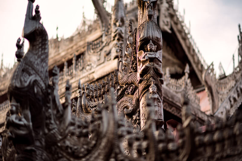 Silver-grey carved teak exterior detail of Shwenandaw Monastery showing celestial figures, nagas, and foliate roof ornament
