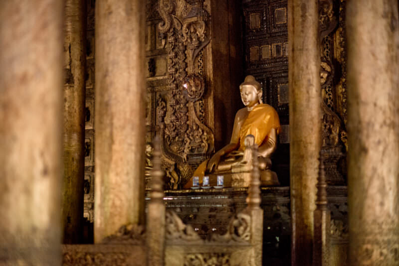Gilded Buddha shrine seen through massive teak columns inside Shwenandaw Monastery, carved posts framing interior space