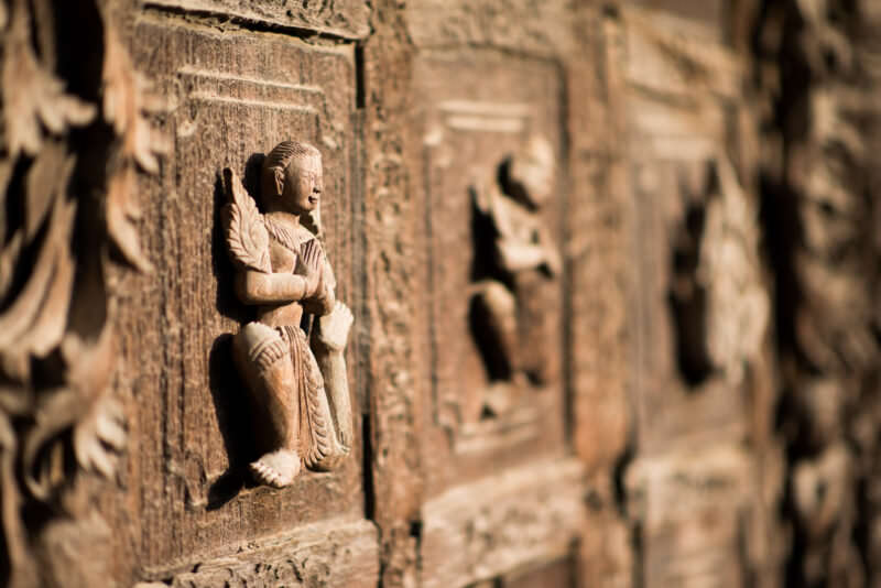 Praying teak figure in shallow relief on weathered exterior panel of Shwenandaw Monastery, gilding entirely stripped