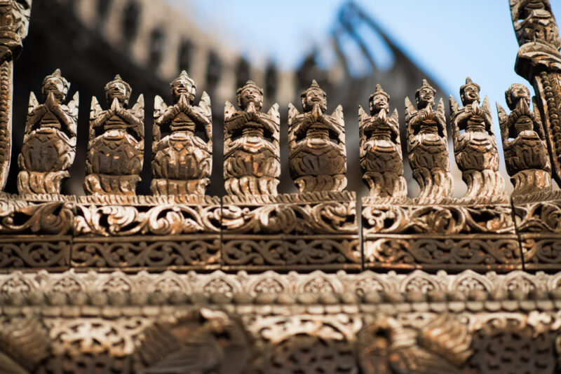 Teak figures in prayer posture lining roofline balustrade of Shwenandaw Monastery above dense foliate relief band