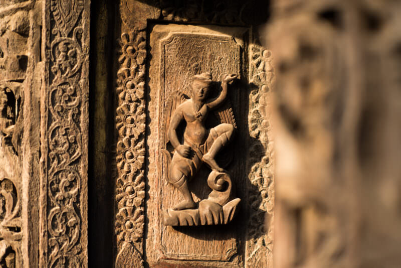 Celestial dancer in high relief on framed teak panel at Shwenandaw Monastery, surrounded by floral scrollwork