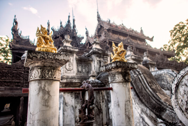 Shwenandaw Monastery courtyard with gilded celestial figures on whitewashed pedestals, weathered teak pyatthat roof rising behind