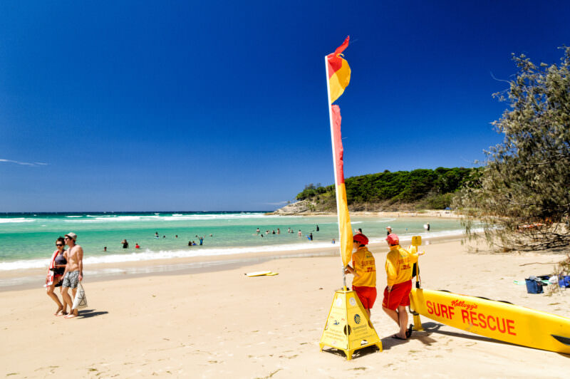 Cylinder Beach on North Stradbroke Island with surf lifesavers in red and yellow beside a tall flag pole and rescue board, swimmers in turquoise water
