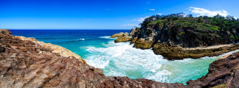 Point Lookout volcanic cliffs with waves surging into a narrow channel, layered rock strata and green-topped headland under blue sky
