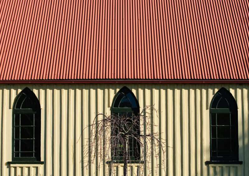 Close-up of a rural church facade showing three Gothic-arched windows in vertical timber siding beneath red corrugated iron roof