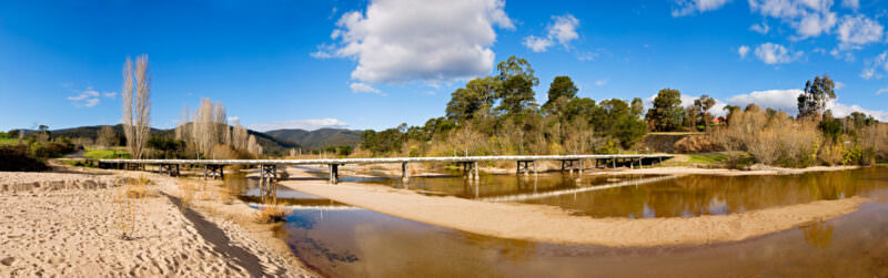 Timber trestle bridge spanning the shallow Towamba River across wide sand banks under blue sky with scattered clouds