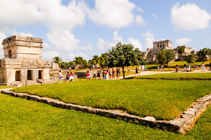 Temple of the Frescoes and other Tulum structures viewed across green lawns under a partly cloudy sky