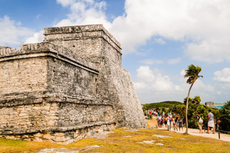 El Castillo pyramid at Tulum, its angled stone walls and flat-topped temple with tourists gathering along the clifftop pathway