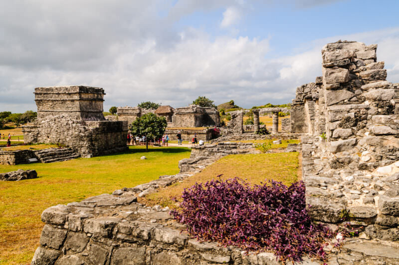 Tulum ruins viewed from within, weathered limestone walls and structures framing green plazas under a cloudy sky