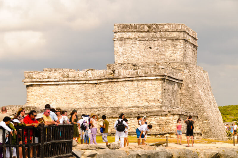 El Castillo at Tulum viewed from the beach, its weathered stone structure seen from sea level with tourists in foreground