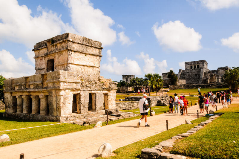Temple of the Frescoes at Tulum, its two-tiered stone structure with columns and dark doorways, surrounded by tourists on pathways