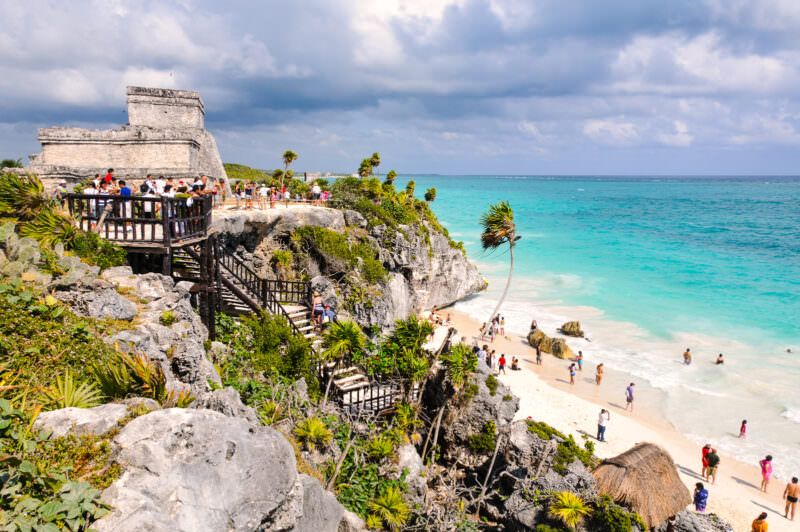 El Castillo at Tulum perched on limestone cliffs above turquoise Caribbean waters, with a wooden viewing platform and visitors