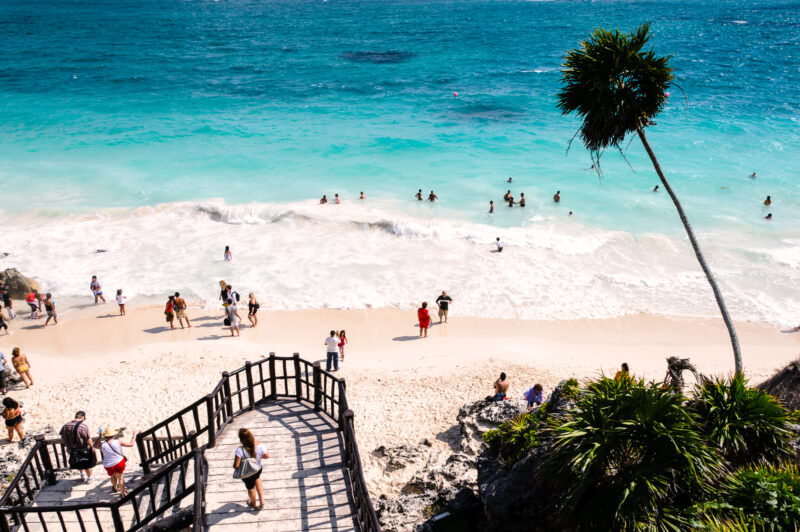 Tulum Beach below the Maya ruins, visitors on white sand and wading in turquoise Caribbean waters with a palm tree in foreground