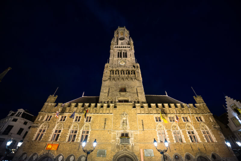 Belfry of Bruges and Hallen illuminated at night, the tower rising above the arched facade on the Markt under deep blue sky