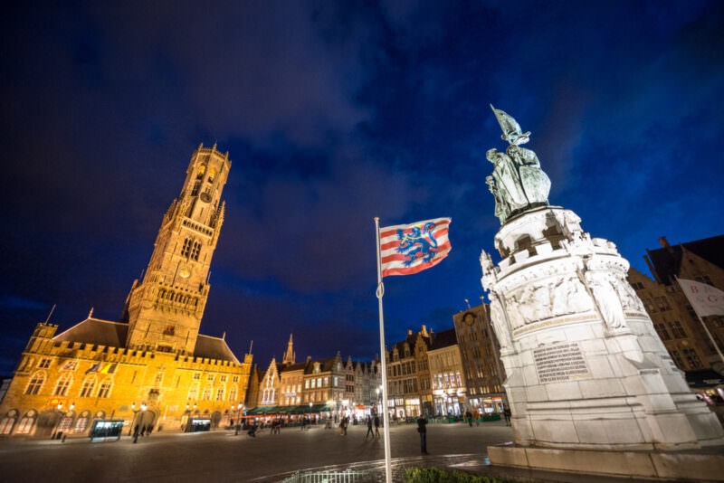 Belfry of Bruges and Breydel-de Coninck monument illuminated at night in the Markt, with West Flemish flag flying beside the bronze statue